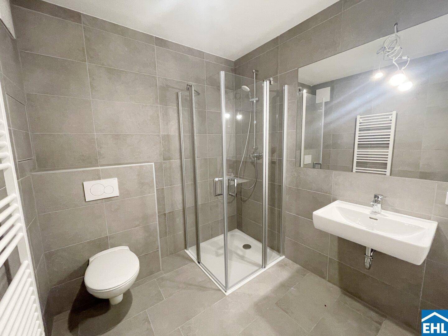 Modernes Badezimmer mit grauen Fliesen, Duschkabine, wandhängendem WC und beheiztem Handtuchhalter.