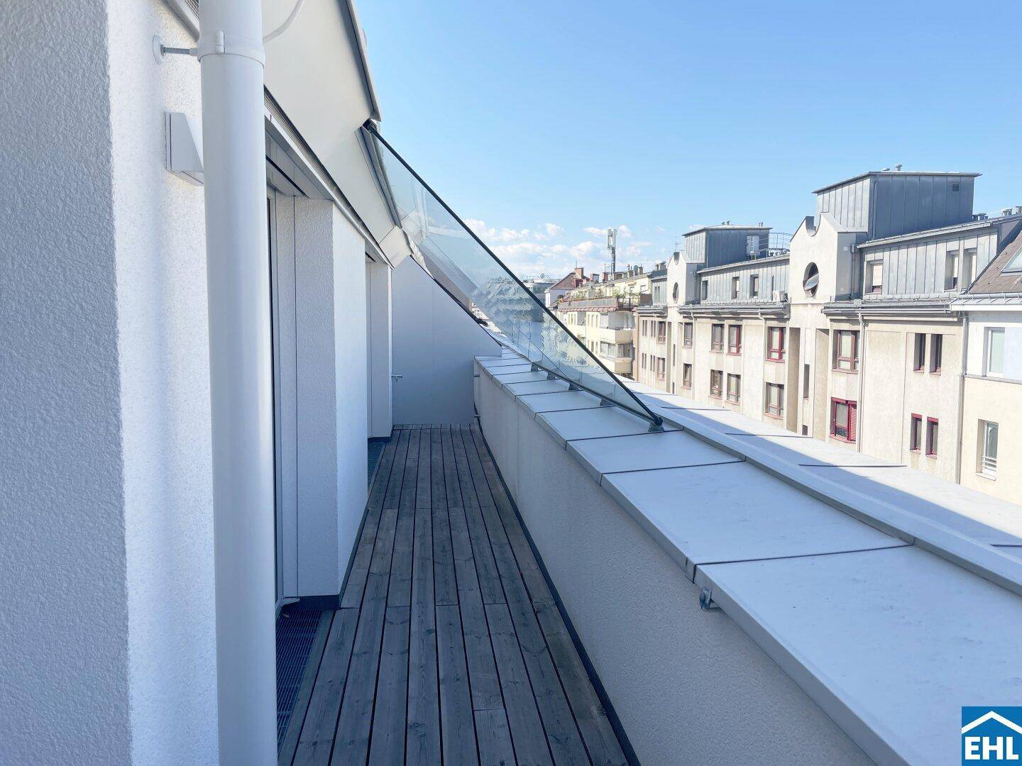 Schmaler Balkon mit Holzboden und Glasgeländer, bietet einen Blick auf die umliegenden Gebäude.