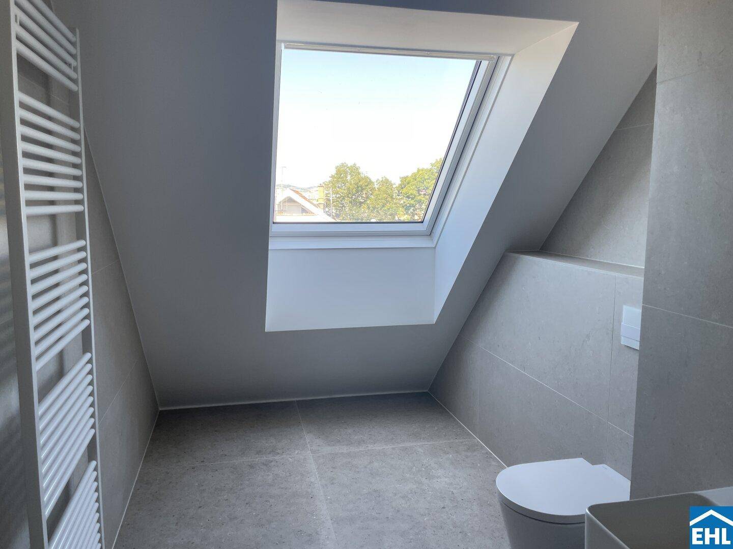 Modernes Badezimmer im Dachgeschoss mit hellgrauen Fliesen, wandhängendem WC und Dachfenster.
