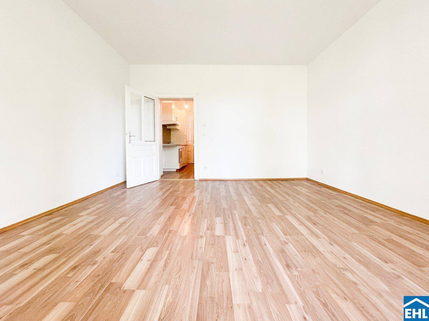 Helles, leeres Zimmer mit Parkettboden und weiß gestrichenen Wänden, mit Zugang zur Küche.