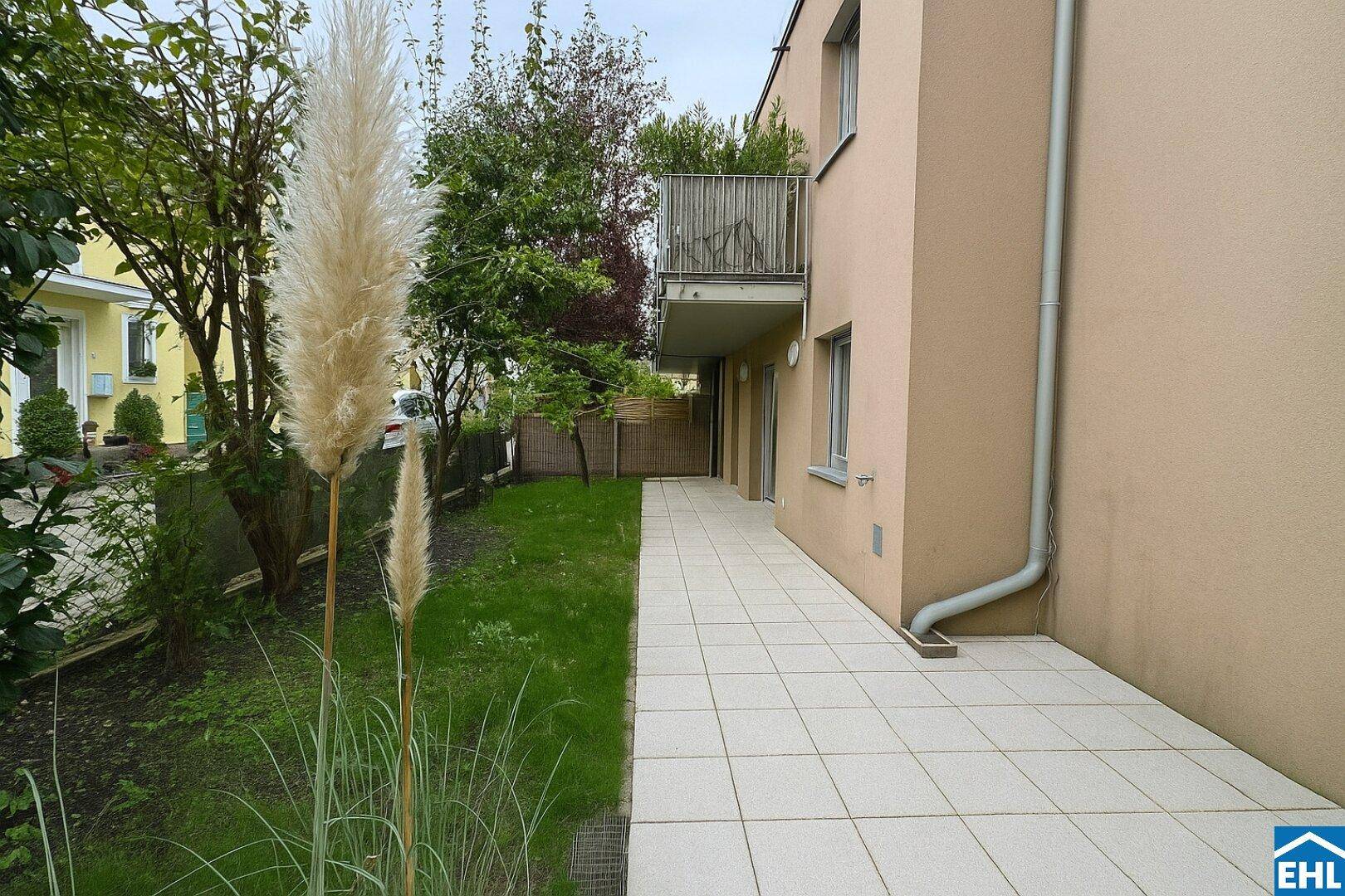 Seitliche Terrasse mit Rasenfläche, Pflanzen und Blick auf den Balkon im Obergeschoss.