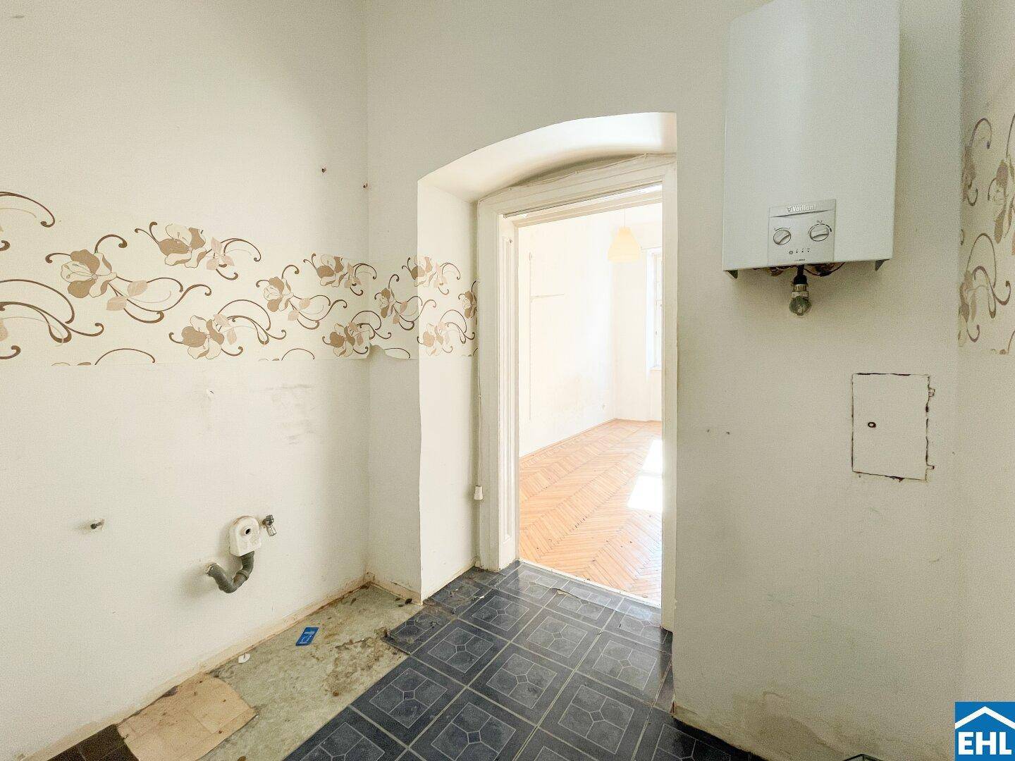 Leere Küche mit Fliesenboden, Gastherme und floraler Tapetenbordüre, renovierungsbedürftig.
