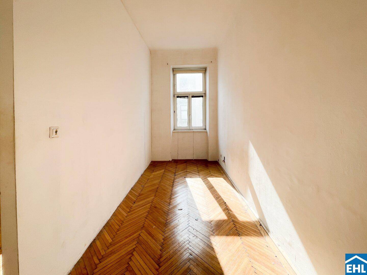 Schmaler, heller Raum mit einem Fenster, Fischgrätparkettboden und natürlichem Lichteinfall.