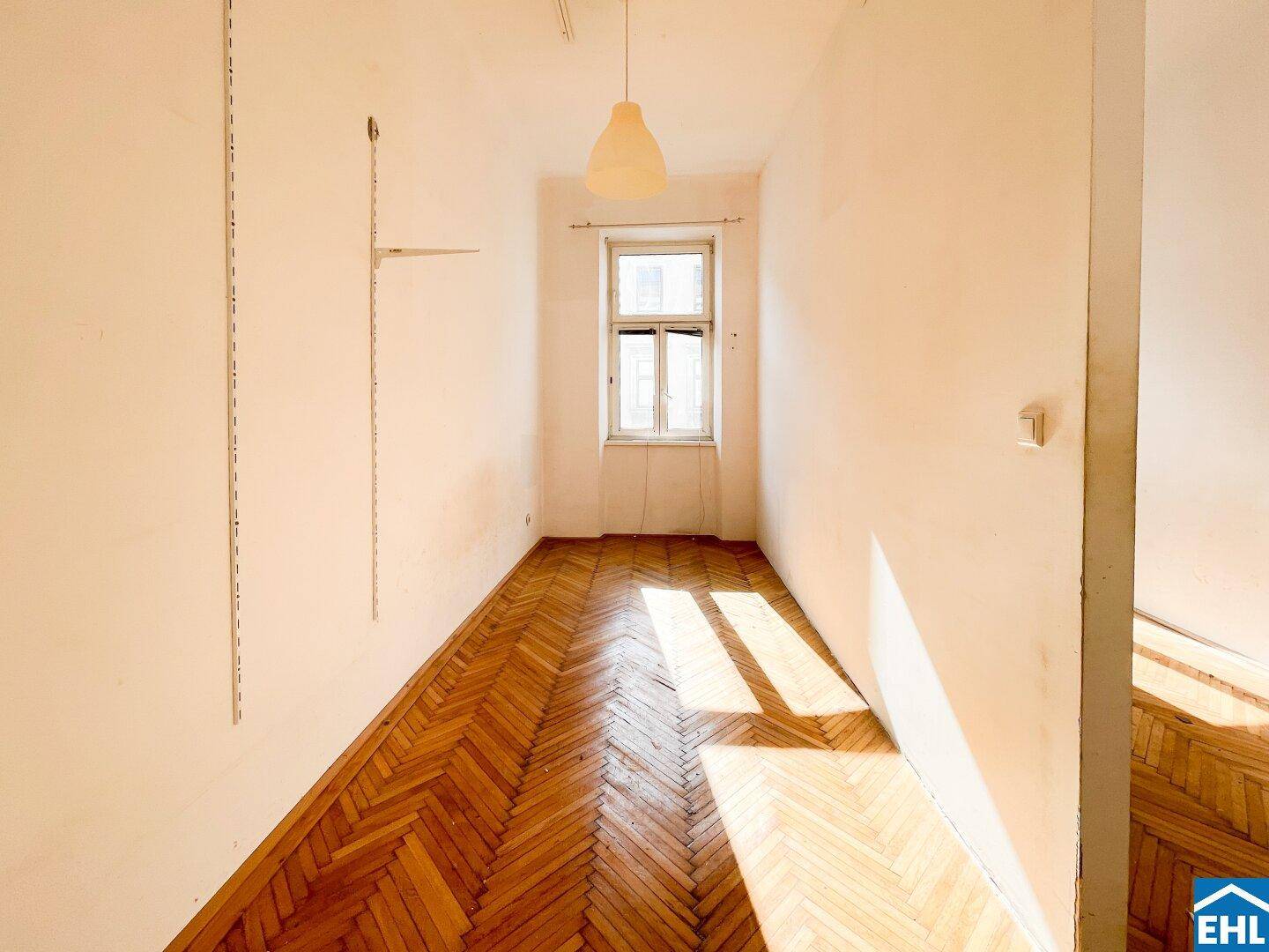 Heller Raum mit Fischgrätparkettboden, einem Fenster und einer einfachen Deckenleuchte.