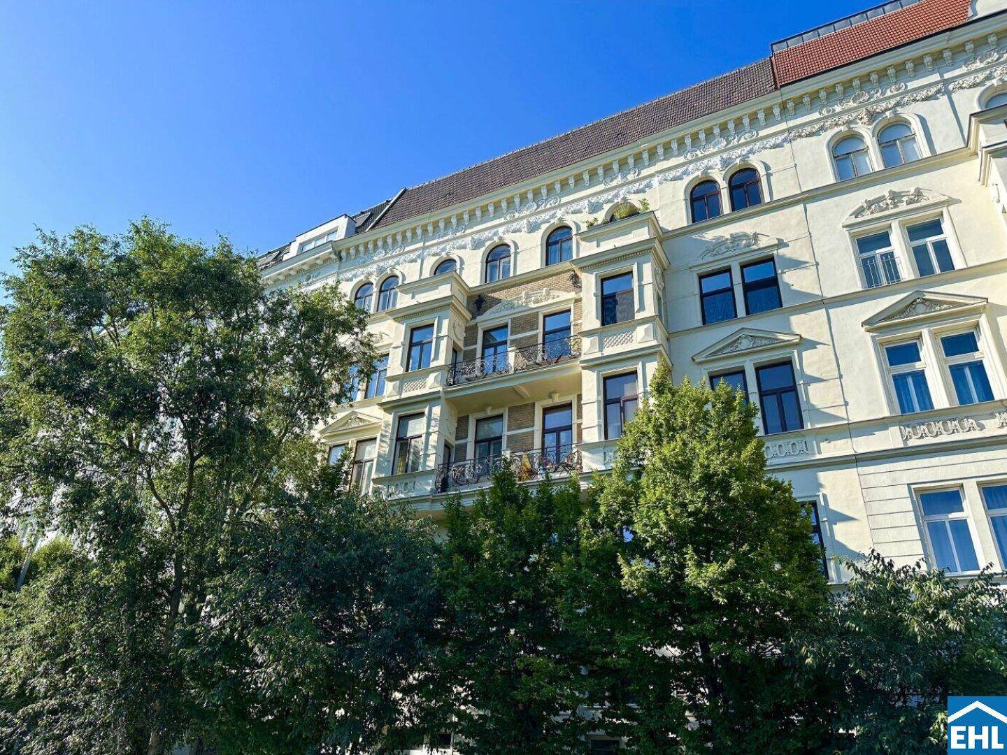 Historische Fassade eines Altbaus mit reich verzierten Details und grünen Bäumen im Vordergrund.