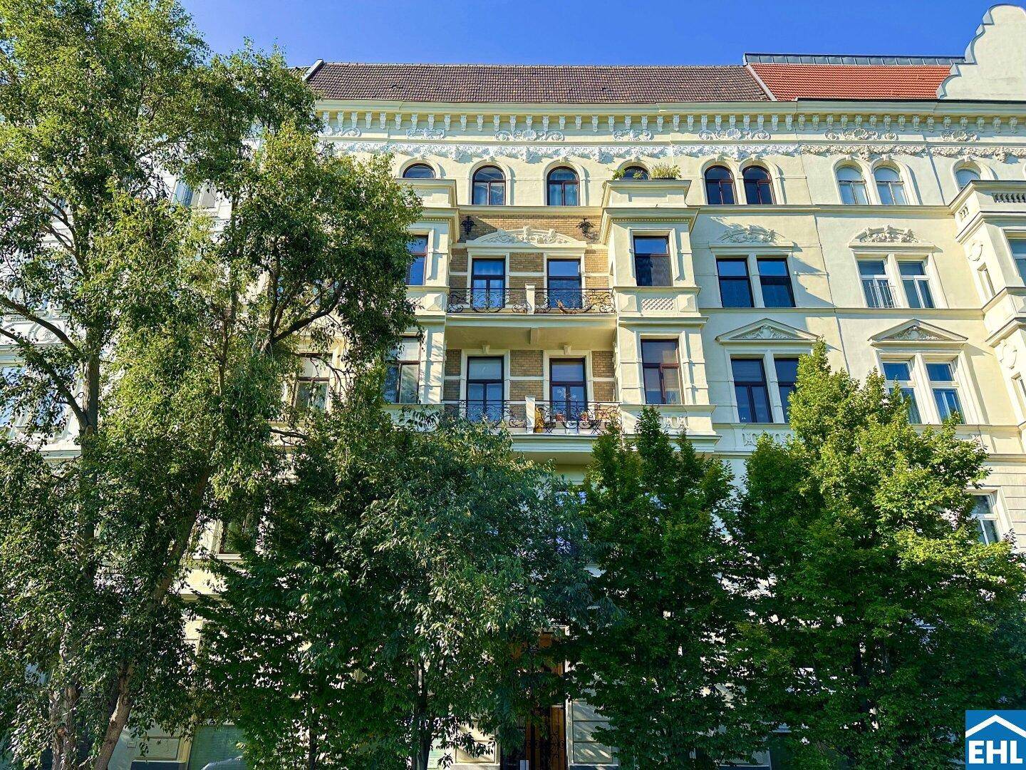 Elegante Altbaufassade mit vielen Fenstern und Balkonen, teilweise von Bäumen verdeckt.
