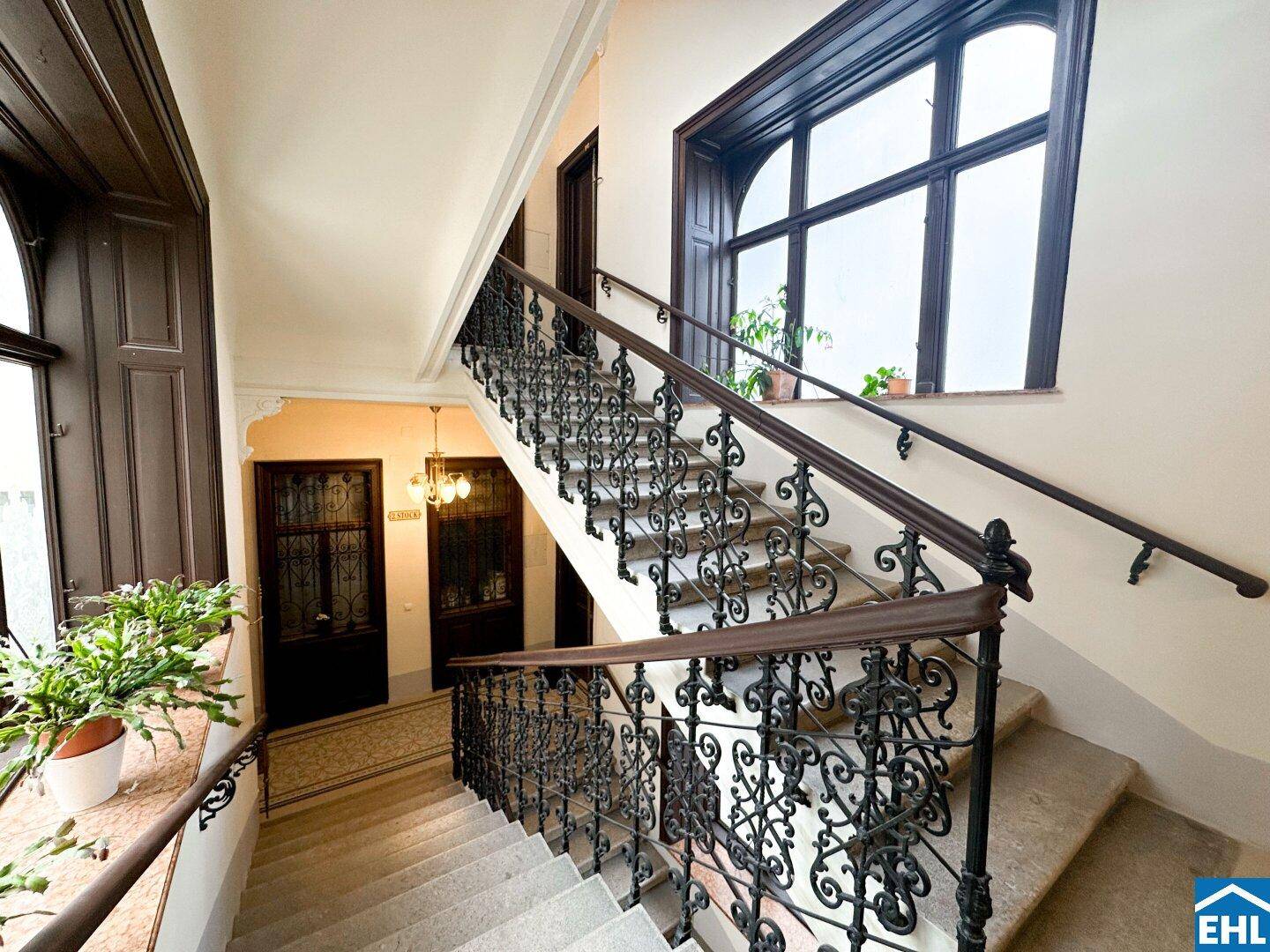Elegantes Treppenhaus eines Altbaus mit schmiedeeisernem Geländer und großen Fenstern.