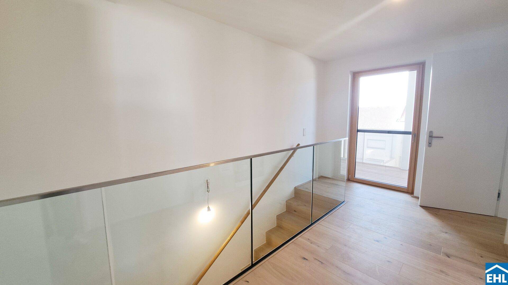 Heller Flur mit moderner Glasbrüstung, Holzboden und Zugang zu einem Außenbereich.