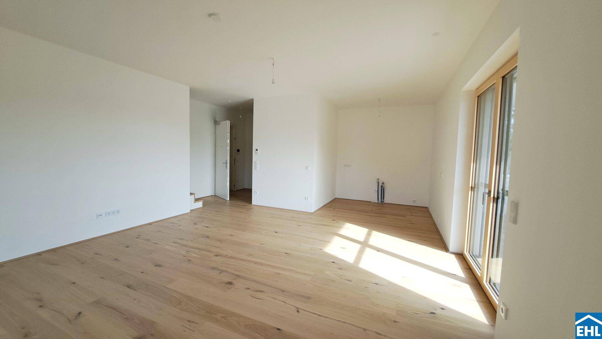 Unmöblierter Wohnraum mit hellem Holzparkett, weißen Wänden und viel Tageslicht durch große Fenster.