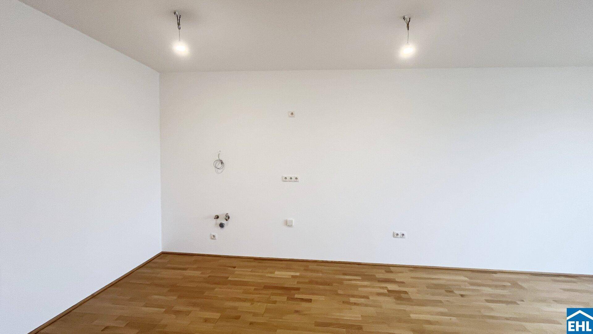 Leerer Raum mit Parkettboden und vorbereiteten Anschlüssen für Medien und Beleuchtung.