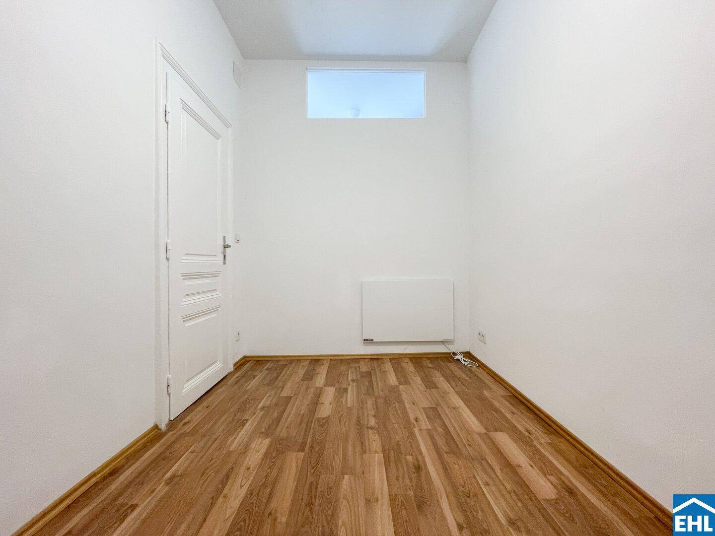 Heller Raum mit Holzboden und einem hohen Fenster, das Tageslicht hereinlässt.