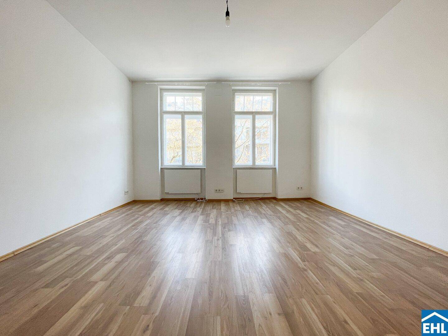 Geräumiges Wohnzimmer mit Holzboden und zwei großen Fenstern, die den Raum erhellen.