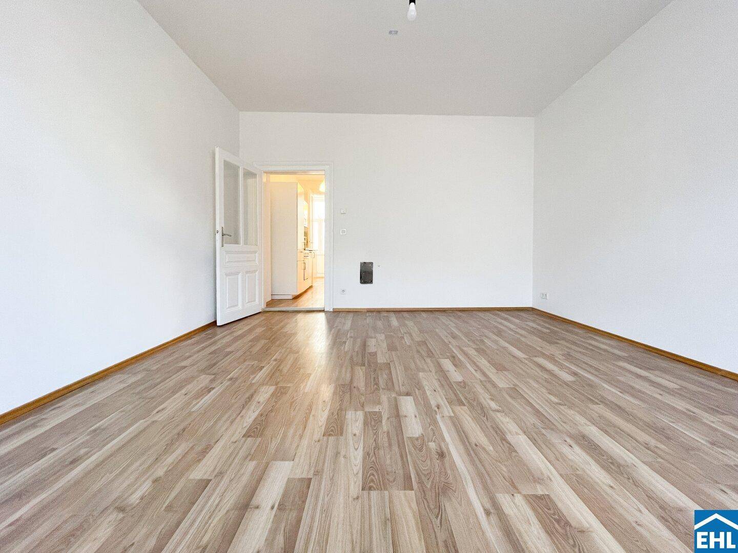 Helles Wohnzimmer mit Holzboden und Zugang zur Küche durch eine offene Tür.