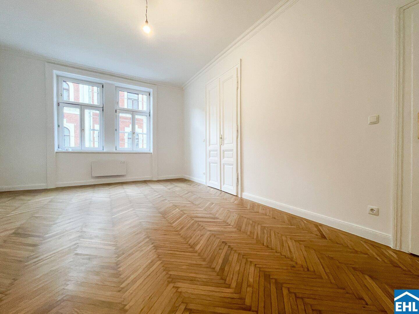 Lichtdurchflutetes Zimmer mit Fischgrätparkett, hohen Fenstern und eleganten Doppeltüren.