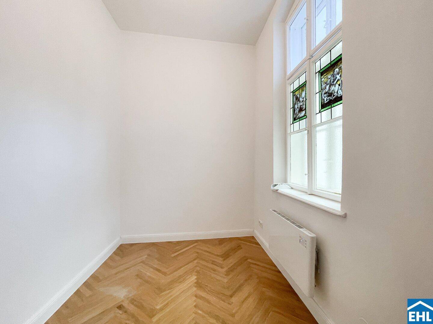 Gemütliches Zimmer mit Fischgrätparkett und einem hohen Fenster mit Buntglas.