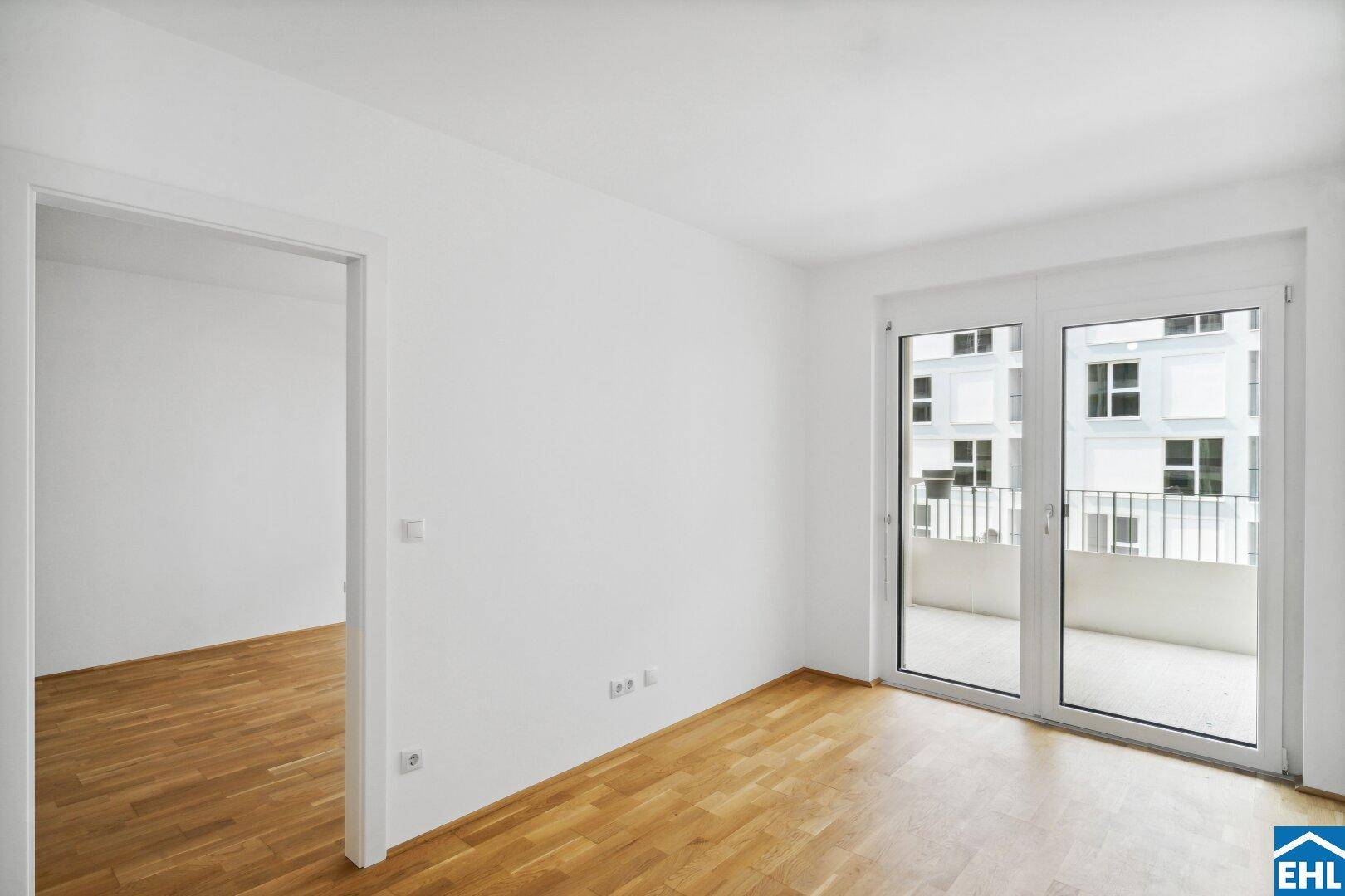 Leerer Wohnraum mit Holzboden, großen Fenstern und Zugang zu einem Balkon.