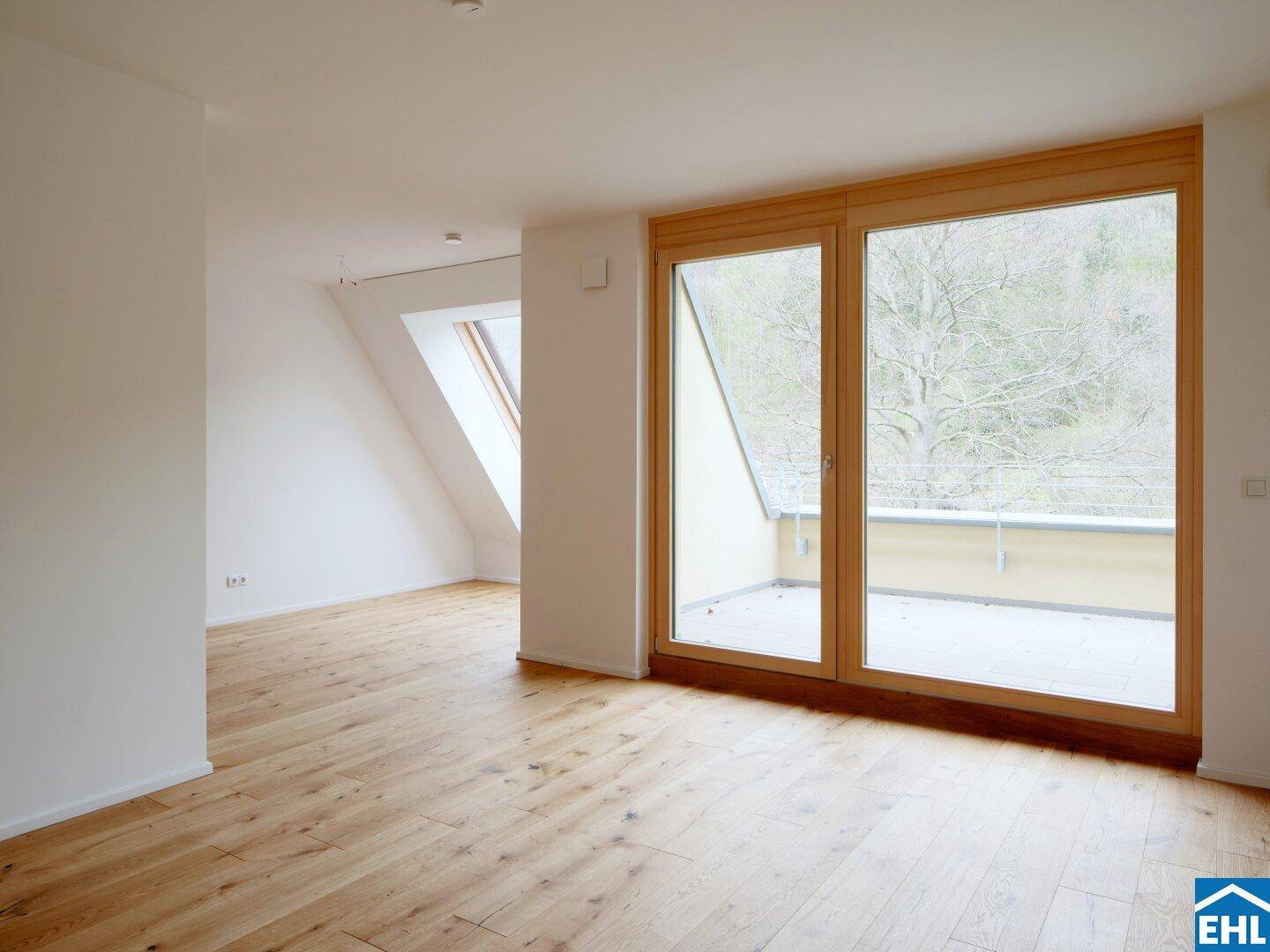 Heller Wohnraum mit Holzboden, großen Fenstern, Dachschräge und Zugang zu einem Balkon.