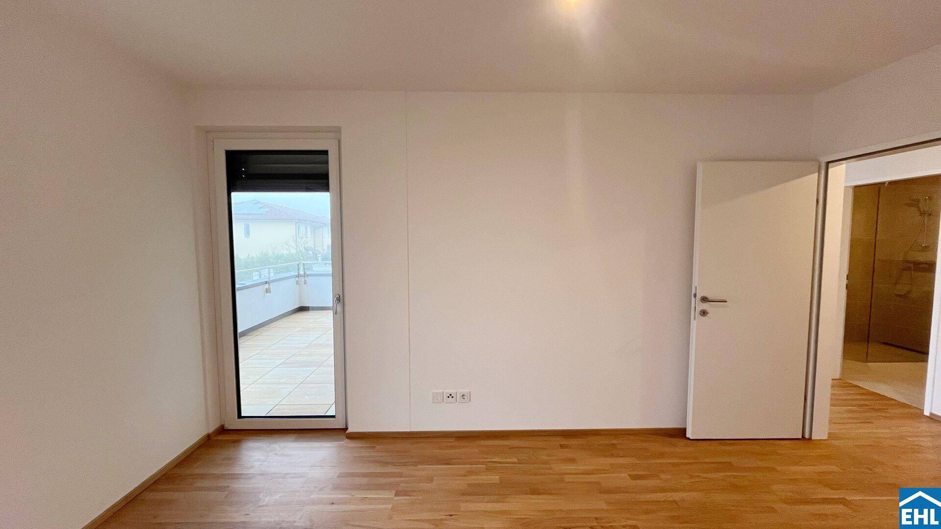 Leeres Schlafzimmer mit Holzboden, Balkonzugang und Tür zu einem angrenzenden Badezimmer.