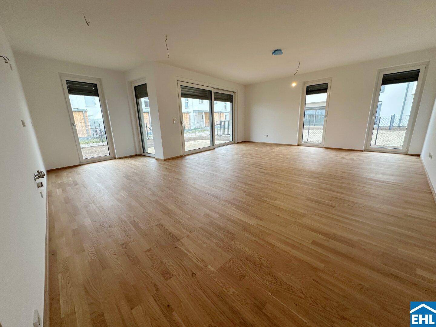 Großer, leerer Wohnbereich mit Parkettboden und mehreren Fenstern, die viel Tageslicht hereinlassen.