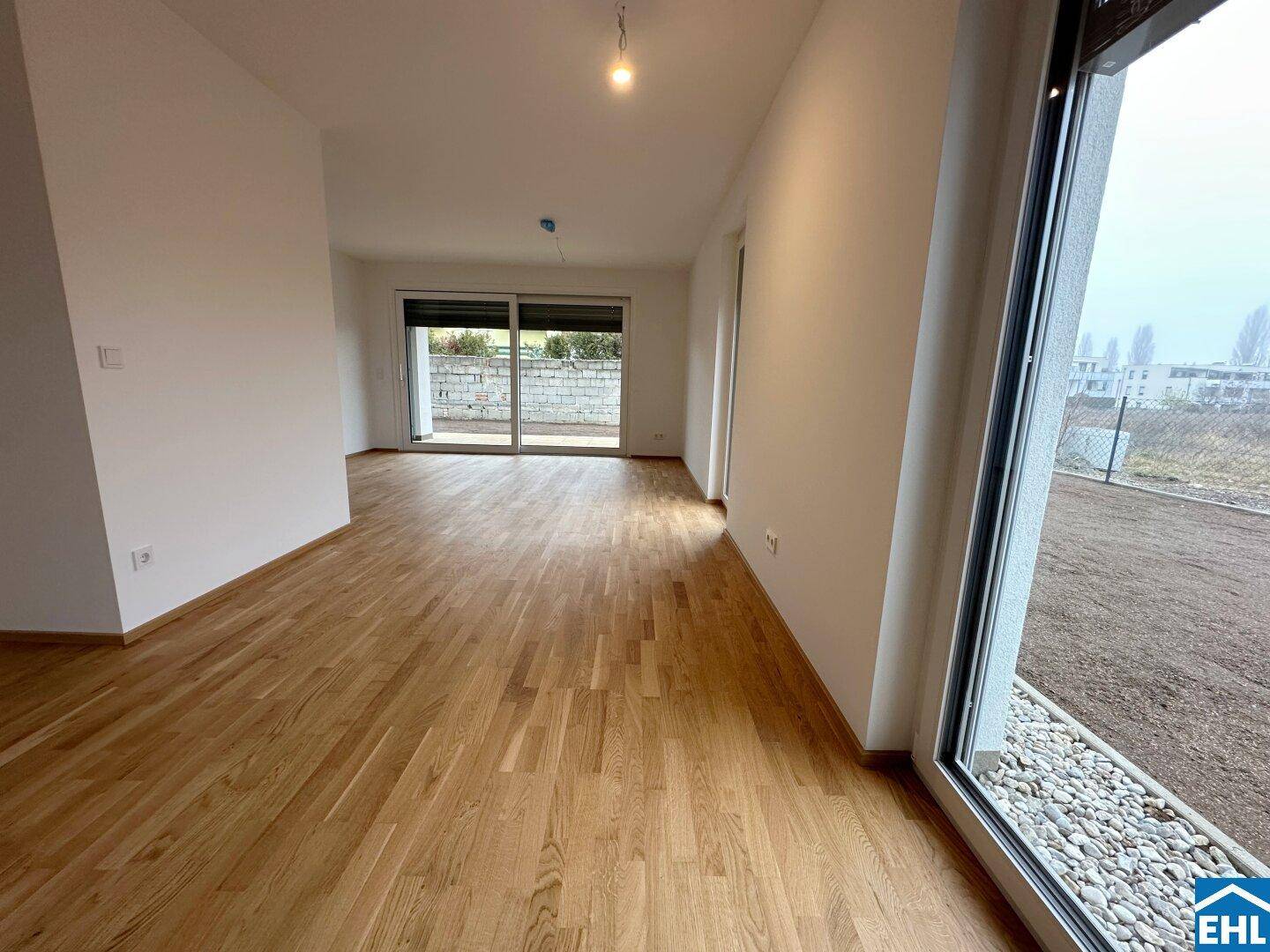 Helles, leeres Wohnzimmer mit Holzboden und großen Fenstern, die viel Tageslicht hereinlassen.