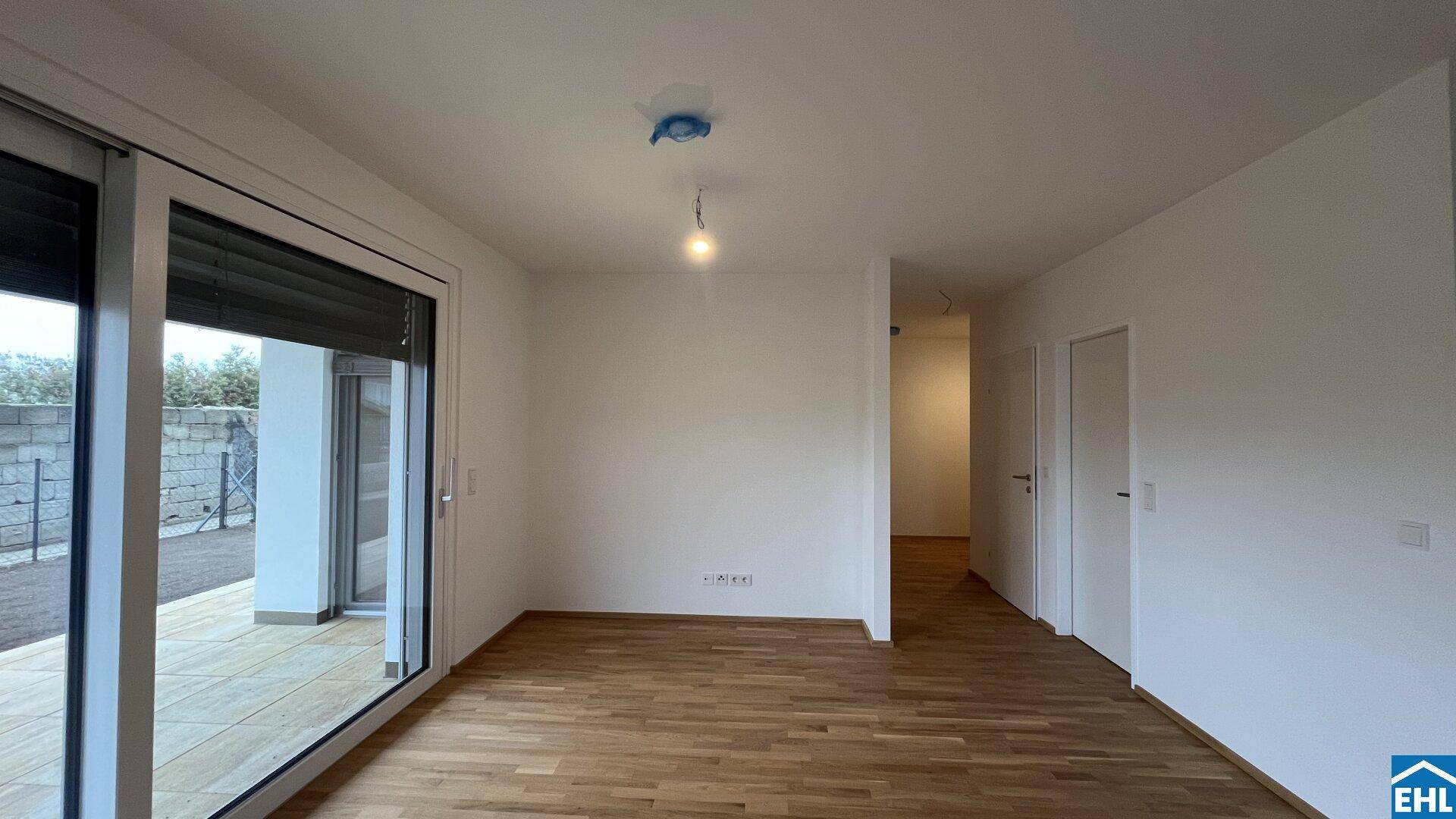 Helles, leeres Wohnzimmer mit Holzboden und großem Fenster zum Außenbereich.