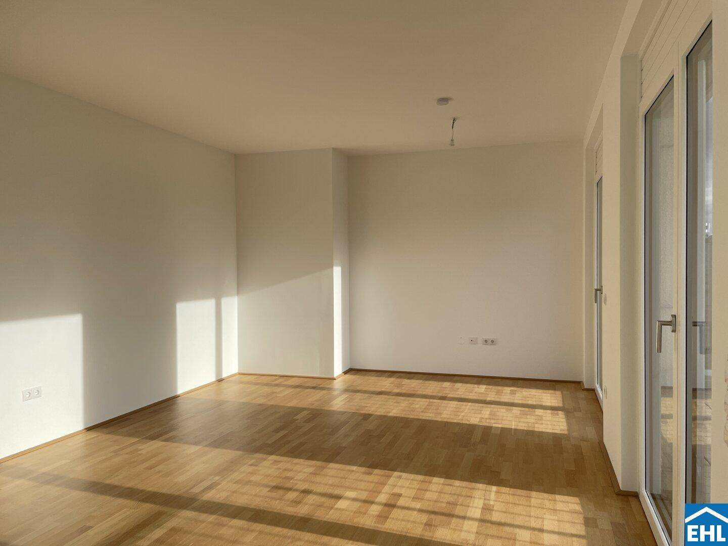Heller, leerer Raum mit Holzboden und großen Fenstern, die viel Tageslicht hereinlassen.