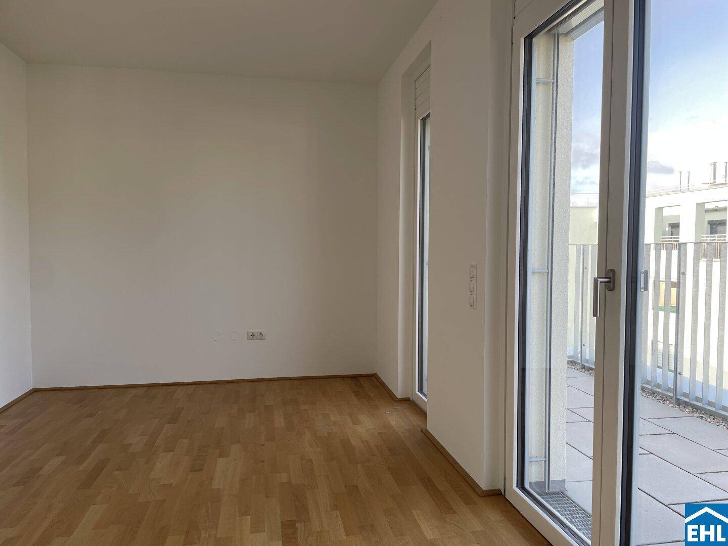 Heller, unmöblierter Raum mit Holzboden und großen Fenstern, die auf einen französischen Balkon führen.