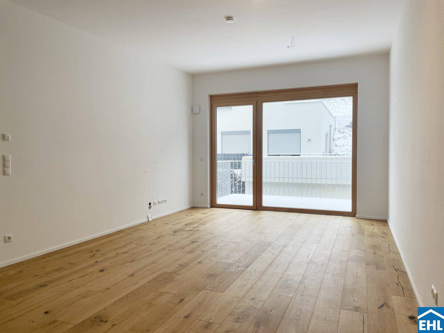 Leeres Wohnzimmer mit hellem Holzboden und großer Glasfront zum Außenbereich.