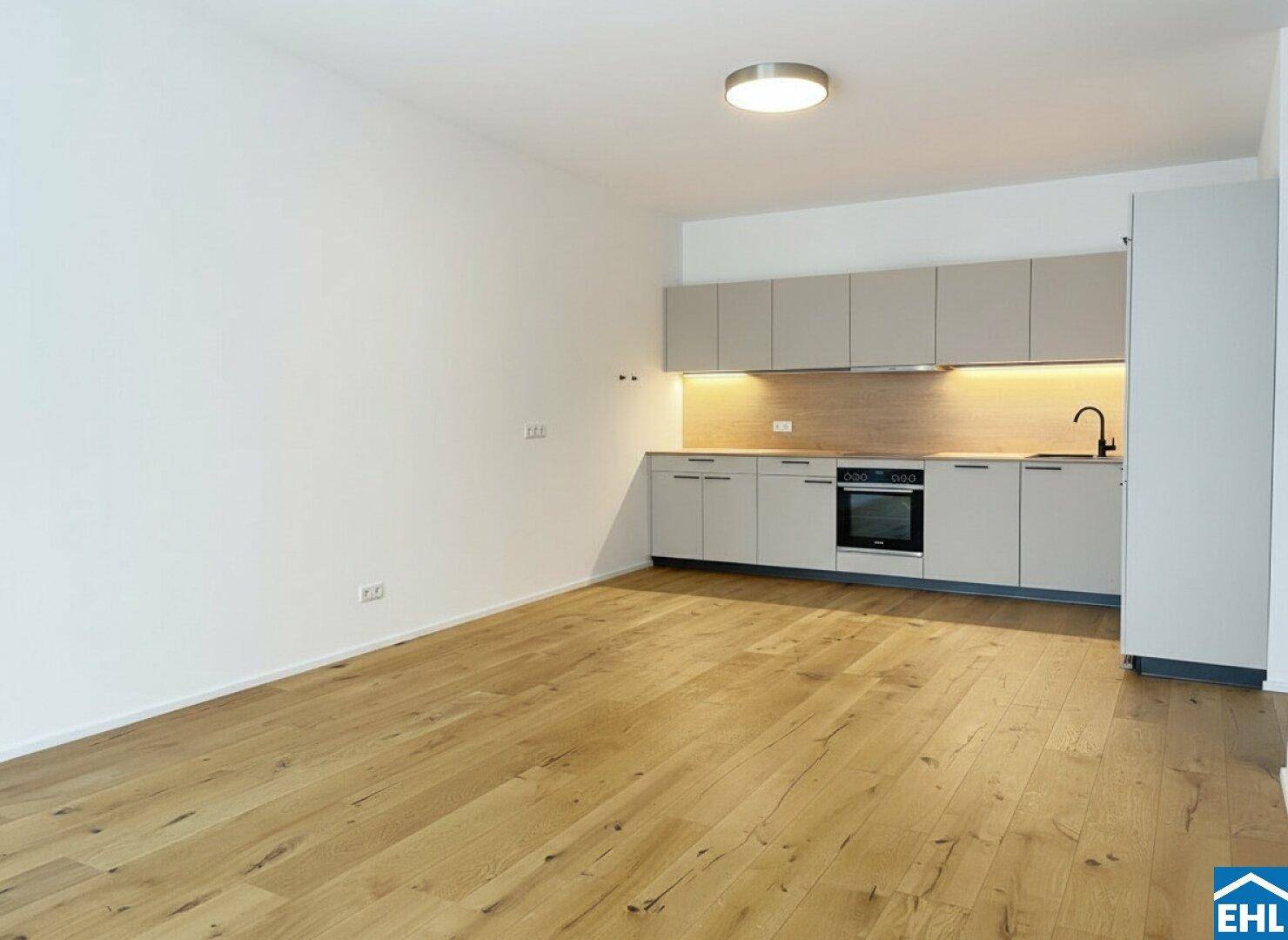 Leerer offener Wohnbereich mit integrierter moderner Küche und hellem Holzboden.