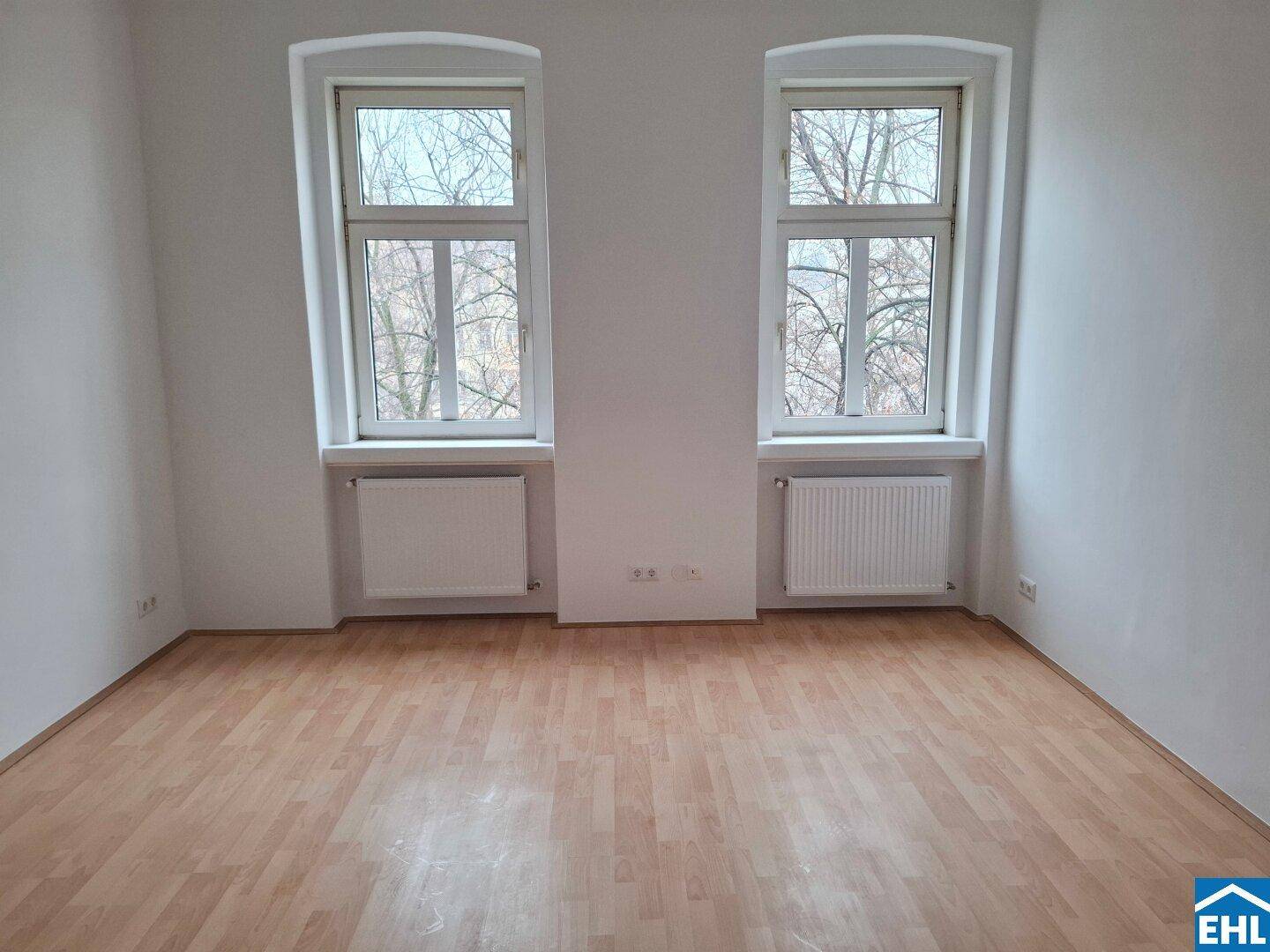 Heller, leerer Raum mit zwei großen Fenstern und hellem Laminatboden.