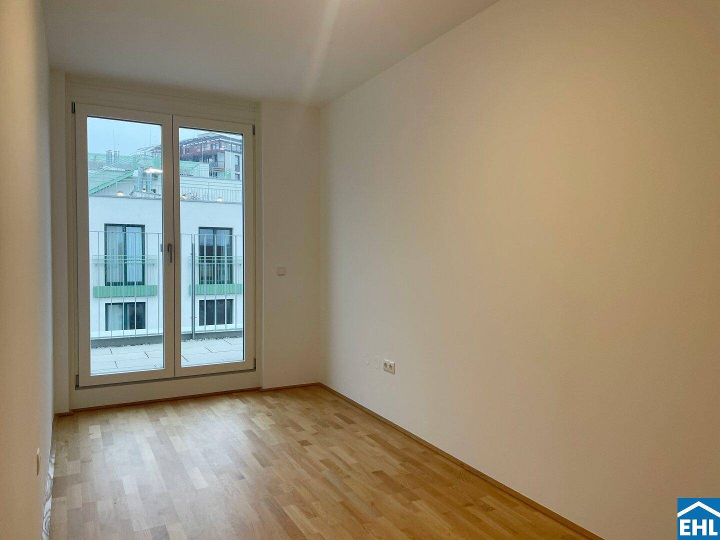 Leeres Zimmer mit Parkettboden und großen Fenstern, die eine helle und freundliche Atmosphäre schaffen.