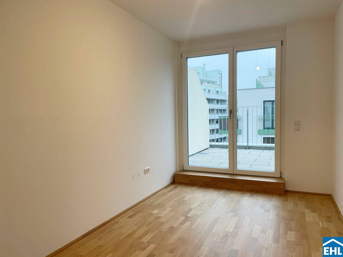 Helles Zimmer mit Parkettboden und großem Fenster, das viel Tageslicht hereinlässt und Zugang zur Terrasse bietet.
