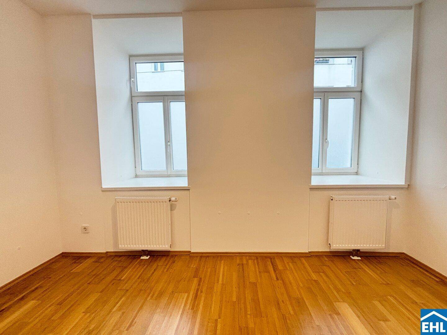 Leeres Zimmer mit Parkettboden und zwei großen Fenstern, bereit zur individuellen Gestaltung.
