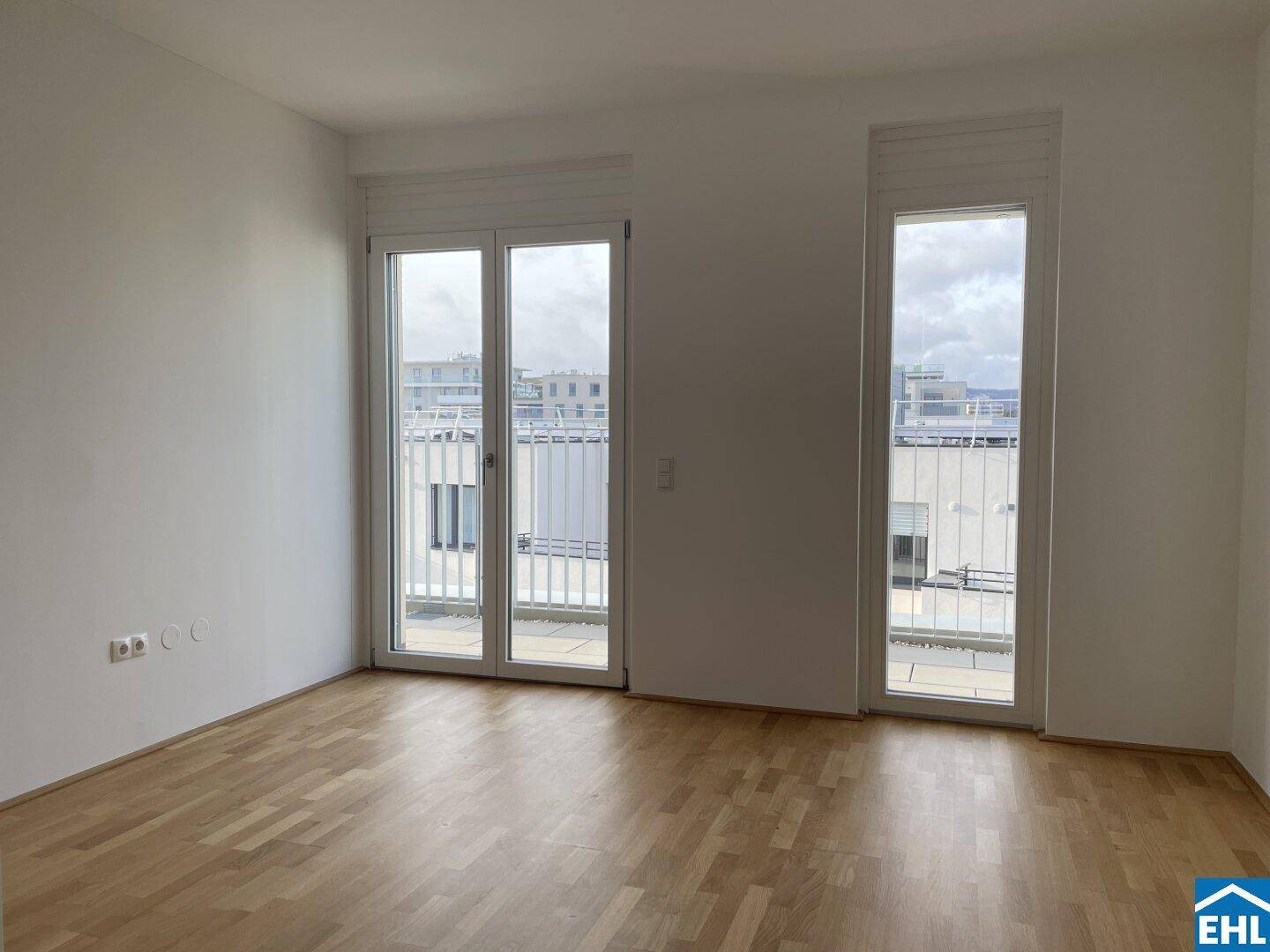 Heller, leerer Raum mit Holzboden und großen Fenstern, die viel Tageslicht hereinlassen.