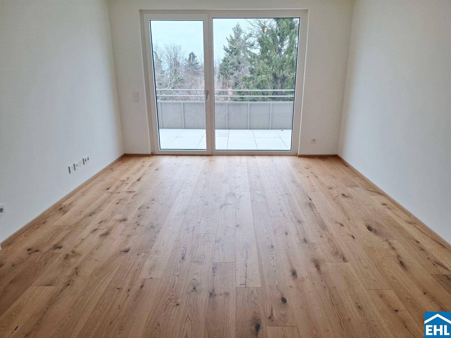 Leerer Raum mit hellem Holzboden und großem Fenster mit Zugang zu einem Balkon.