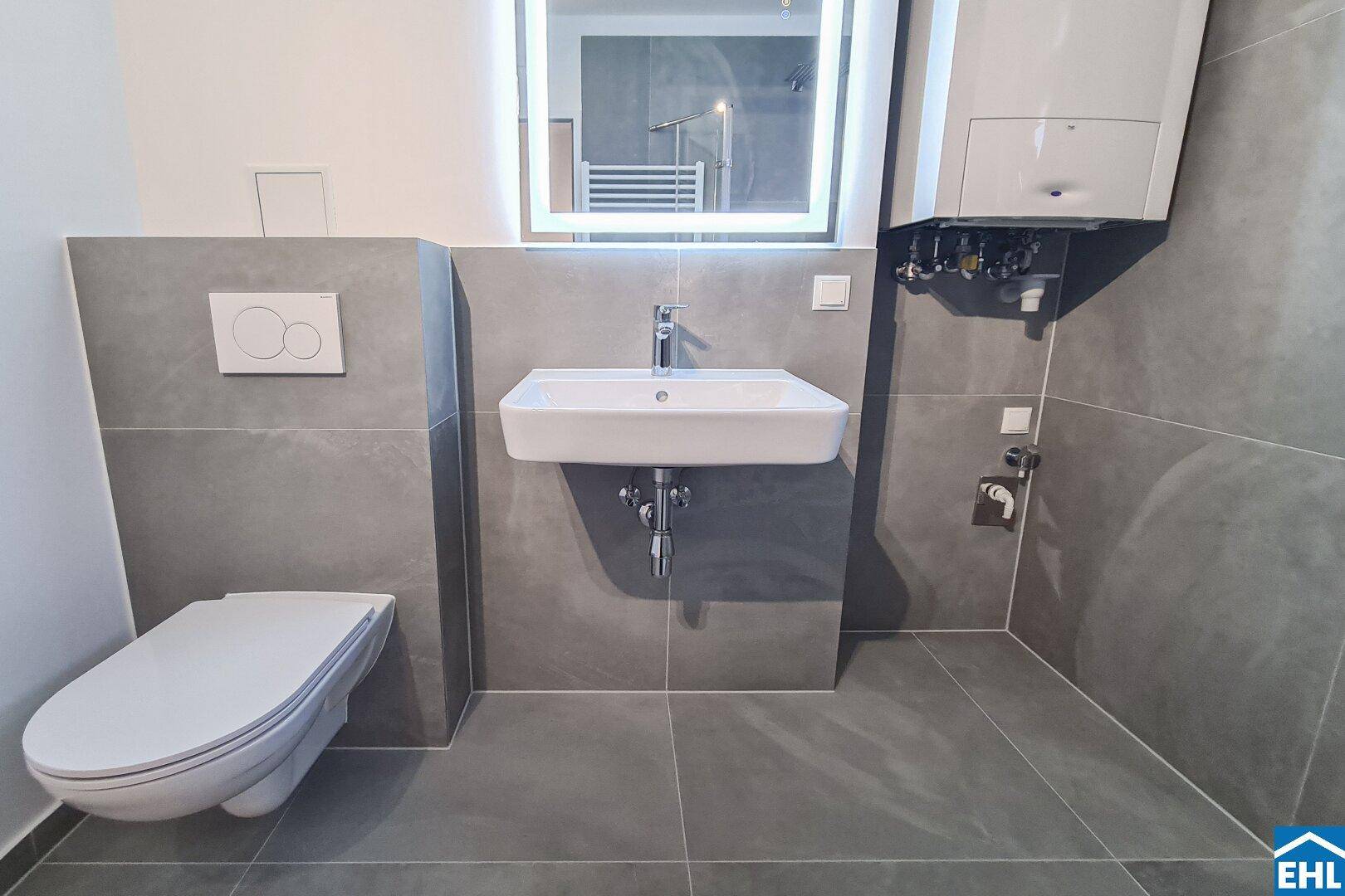 Modernes Badezimmer mit grauen Fliesen, wandhängendem WC und beleuchtetem Spiegel über dem Waschbecken.