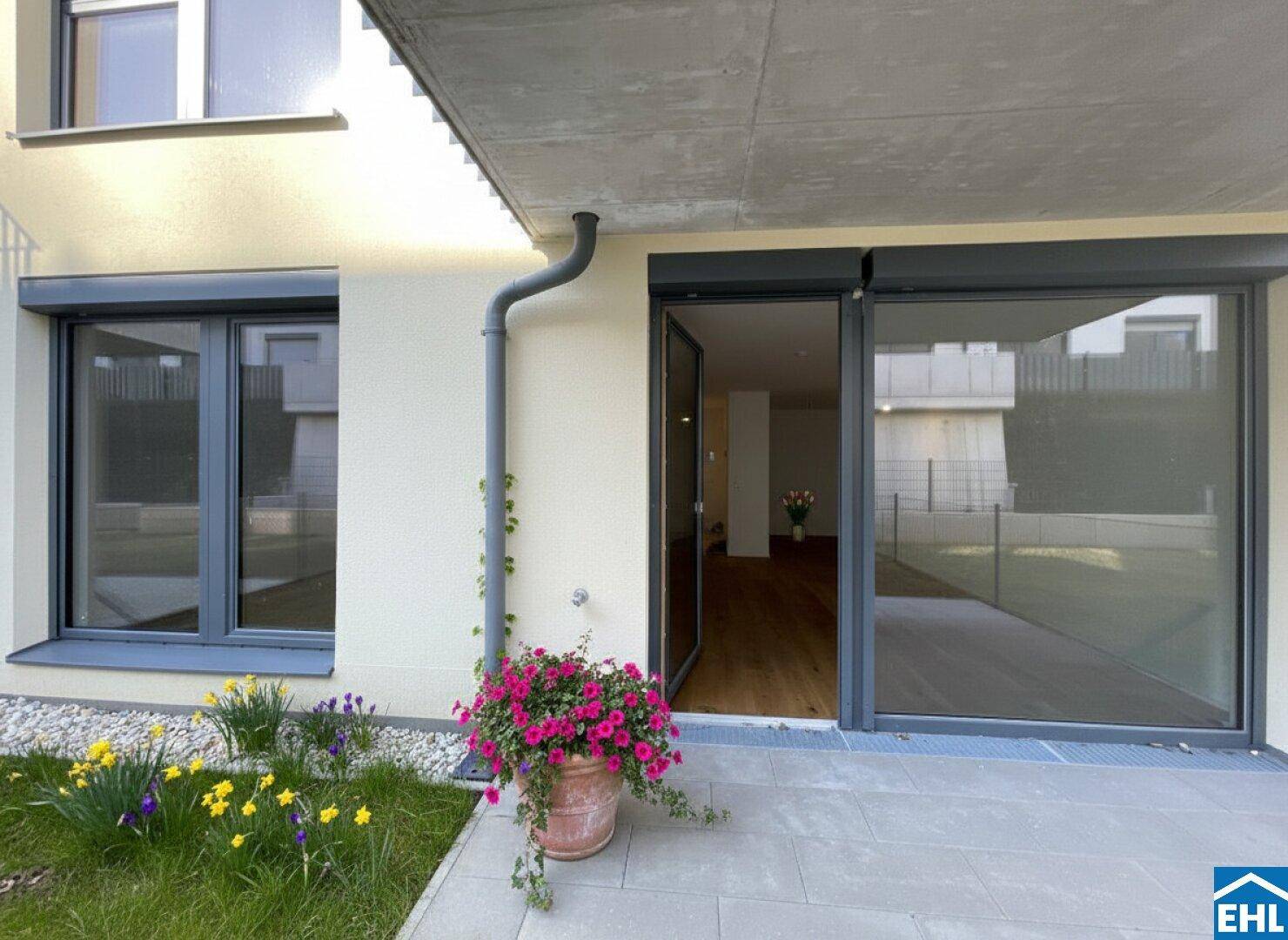 Eingangsbereich einer Erdgeschosswohnung mit kleiner Terrasse, Blumen und großen Fenstern.