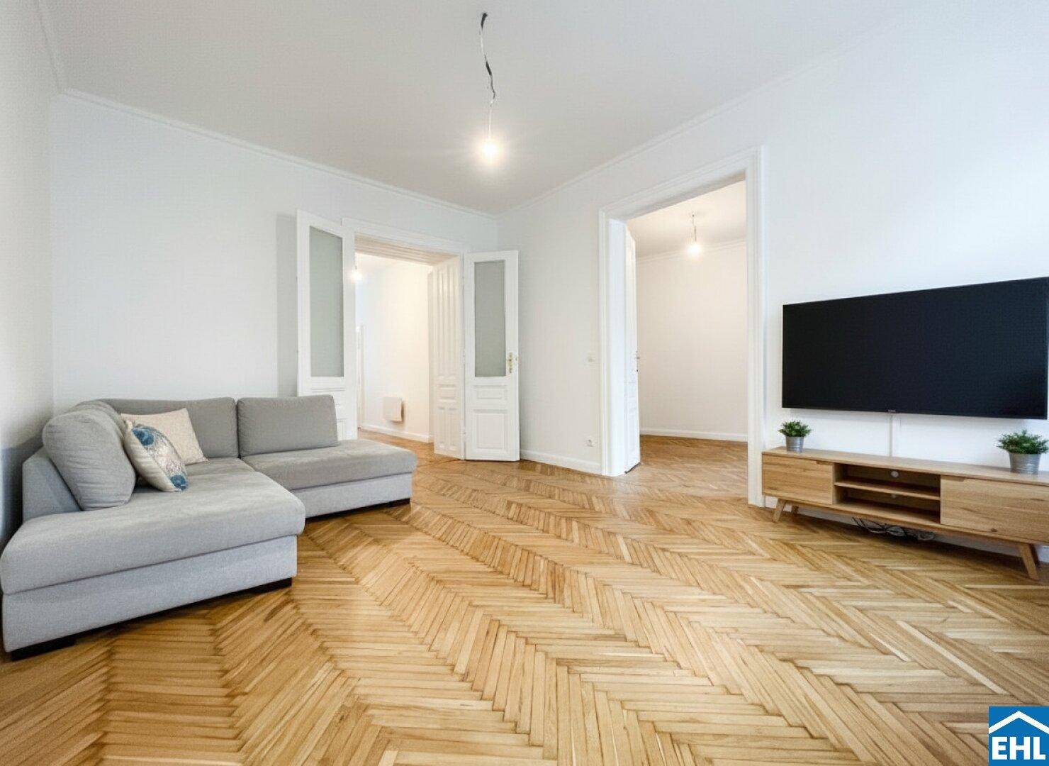 Geräumiges Wohnzimmer mit hellem Fischgrätparkett, modernem Sofa und Fernseher.
