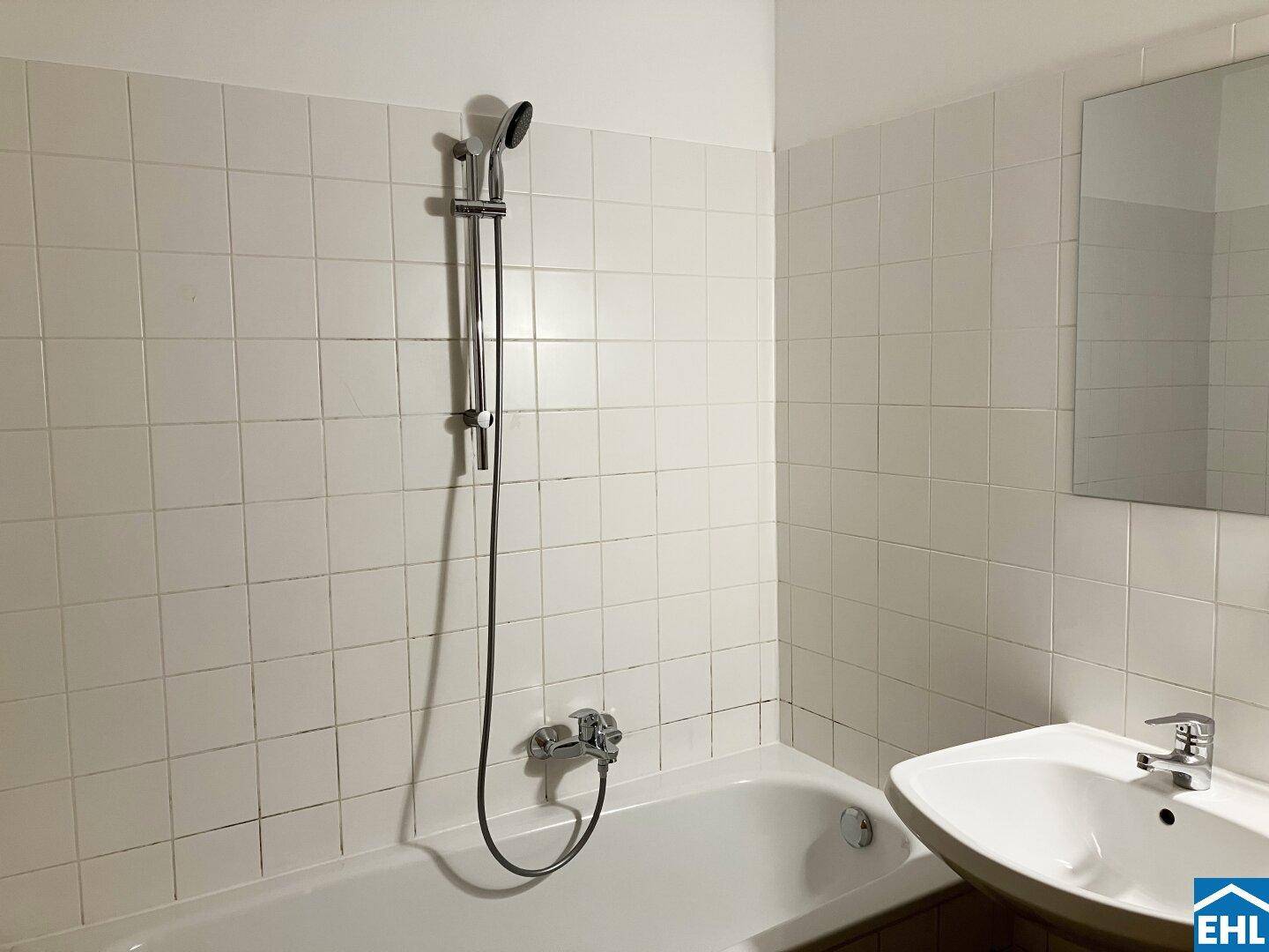Badezimmer mit weißen Fliesen, Badewanne mit Duschfunktion und Waschbecken mit Spiegel, in Standardausstattung.