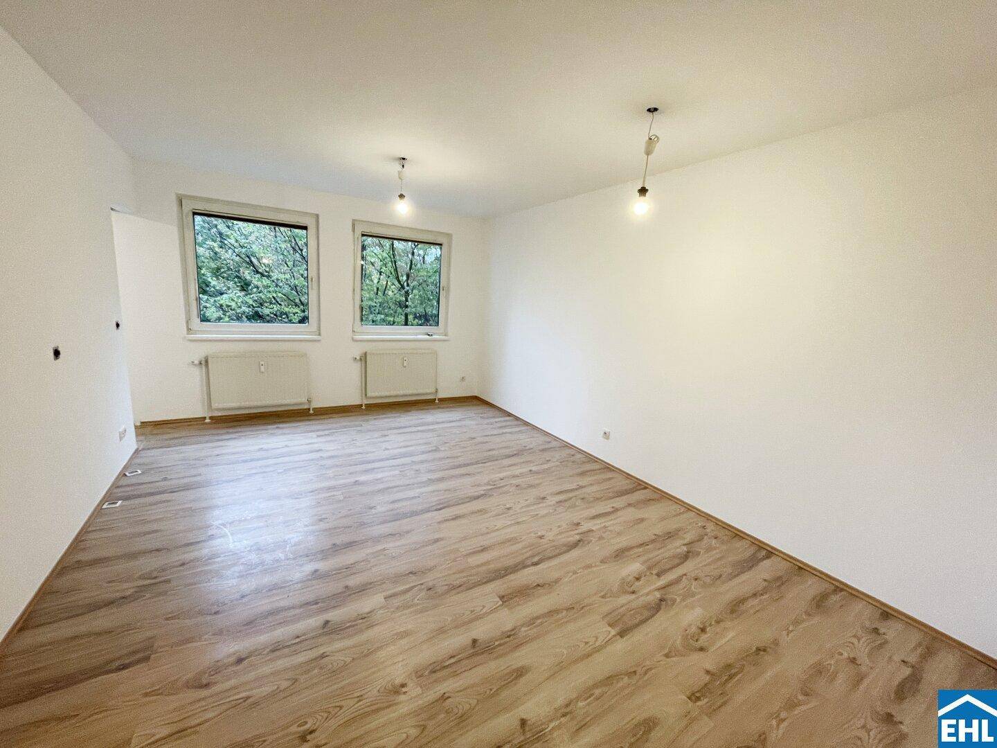 Geräumiges Wohnzimmer mit zwei Fenstern, die einen Blick ins Grüne bieten, und hellem Laminatboden.