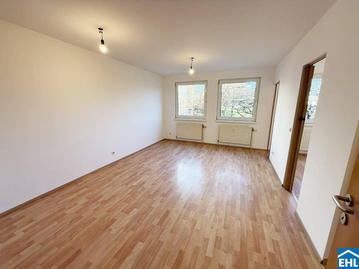 Helles Wohnzimmer mit zwei Fenstern, Laminatboden und weiß gestrichenen Wänden.