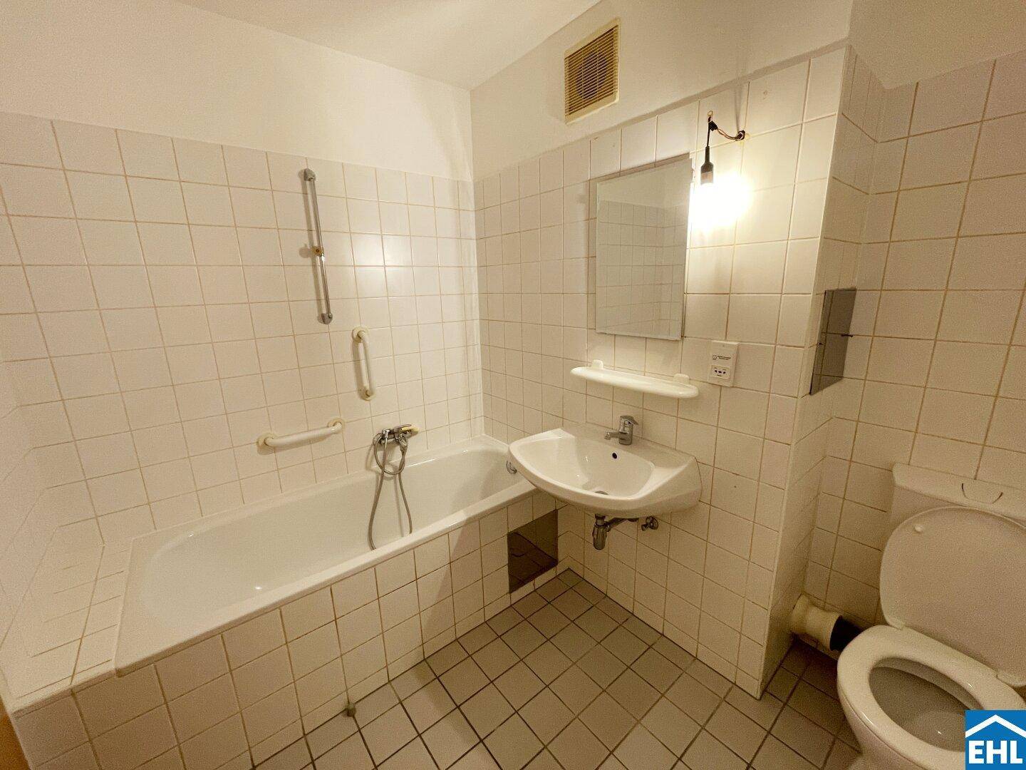 Gefliestes Badezimmer mit Badewanne, Waschbecken und WC, ausgestattet mit Haltegriffen.