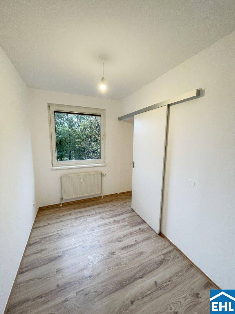 Helles Schlafzimmer mit Laminatboden, Schiebetür und Fenster mit Blick ins Grüne.