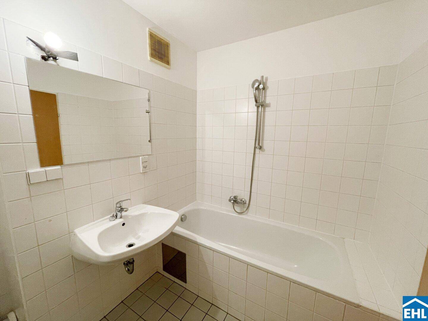 Badezimmer mit weißen Fliesen, Badewanne mit Duschfunktion und Waschbecken mit Spiegel.