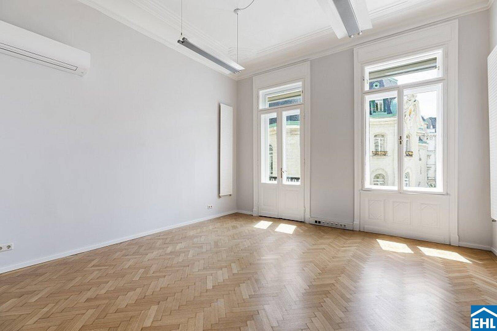 Geräumiger, unmöblierter Raum mit Fischgrätparkett, hohen Decken und zwei großen Fenstern.