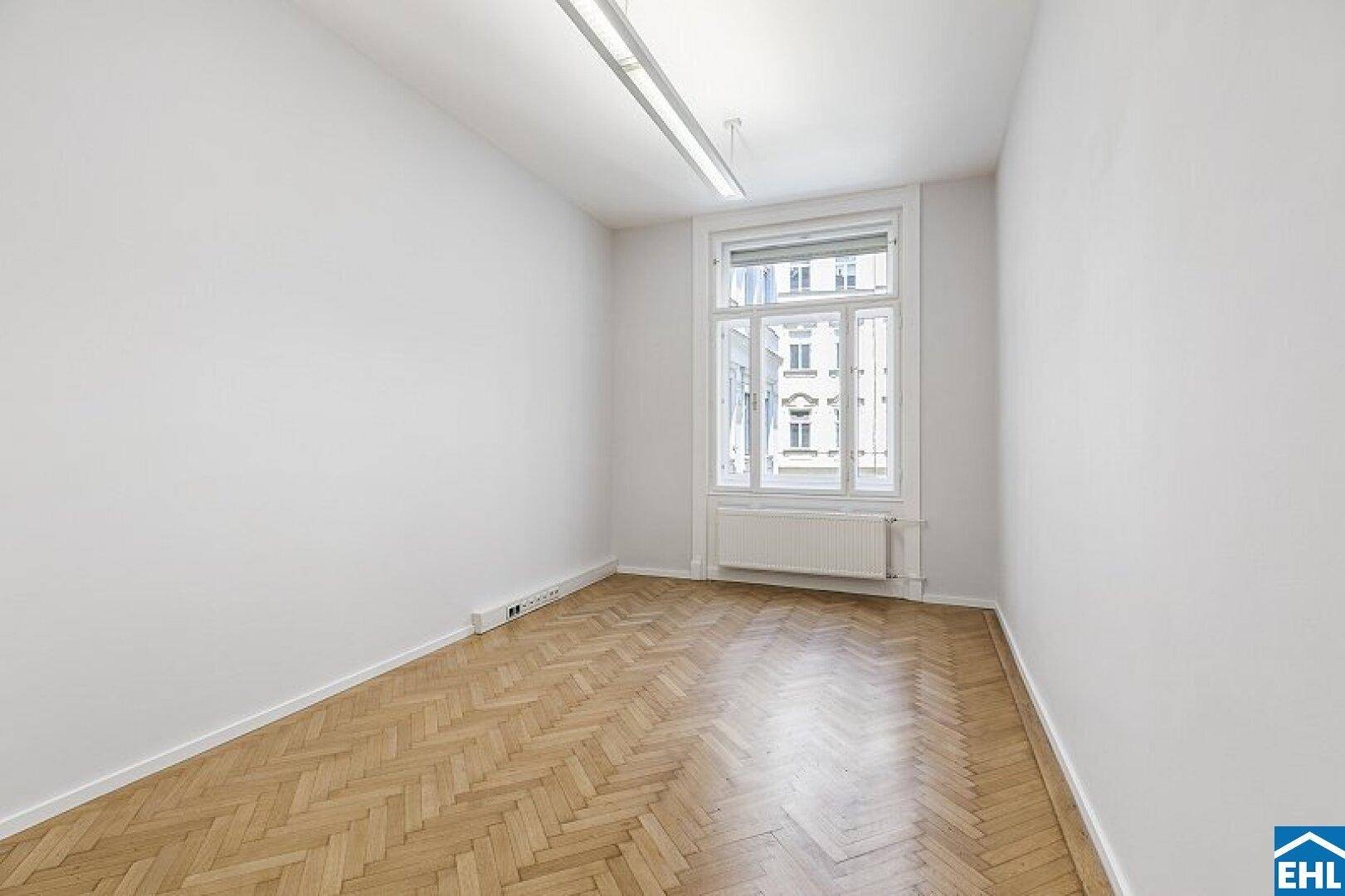 Leerer Raum mit Fischgrätparkett, weißen Wänden und einem Fenster mit Stadtblick.