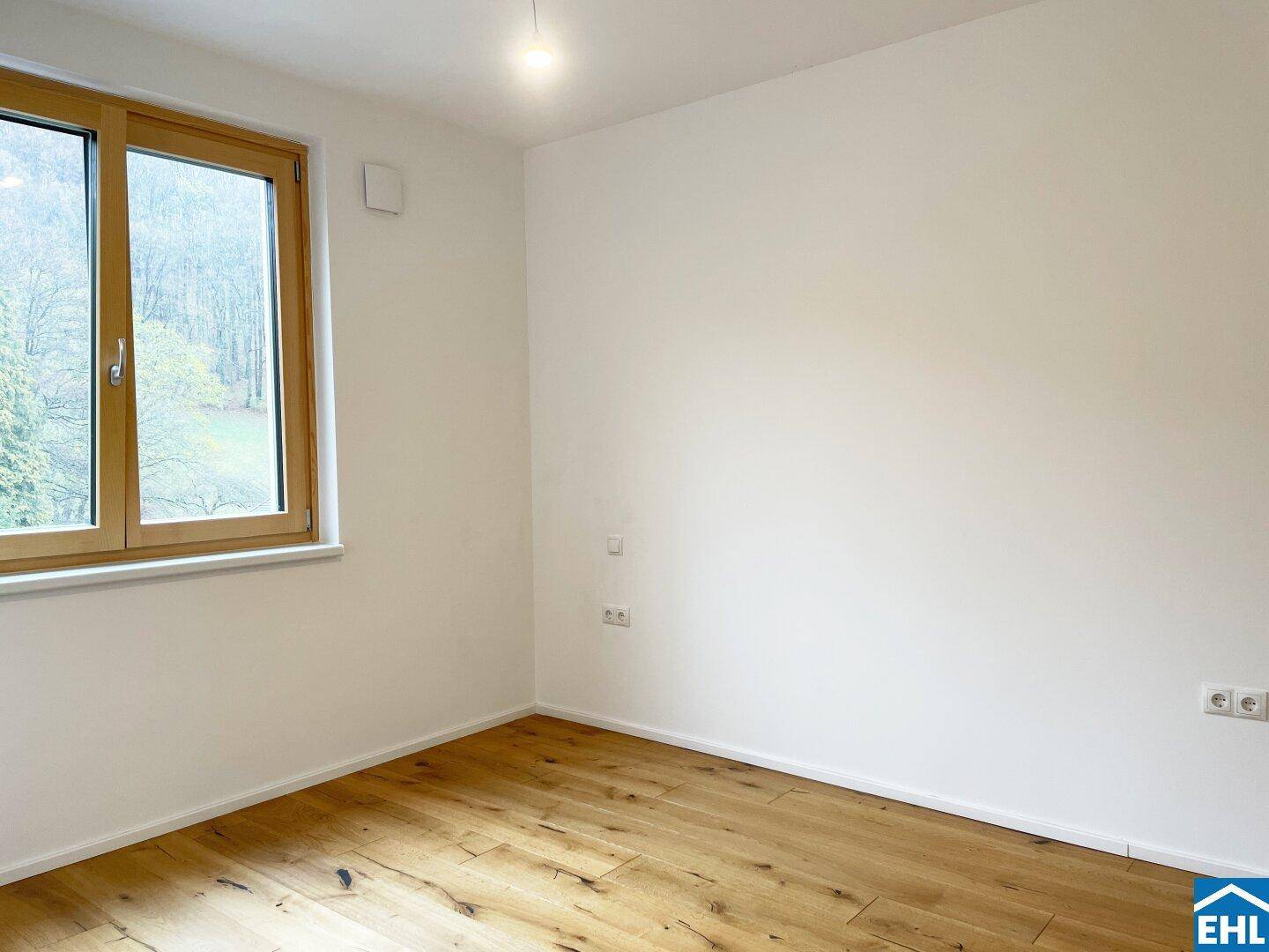 Helles Zimmer mit Holzboden und einem Fenster, das eine angenehme Aussicht ins Grüne ermöglicht.