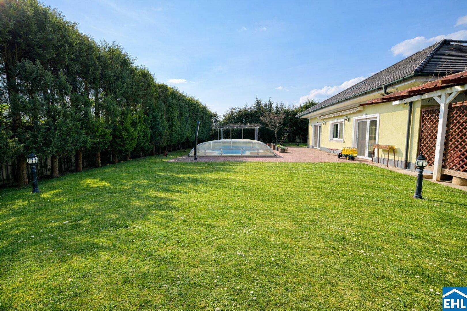 Weitläufige, gepflegte Gartenanlage mit Rasenfläche und Pool im Hintergrund unter blauem Himmel.