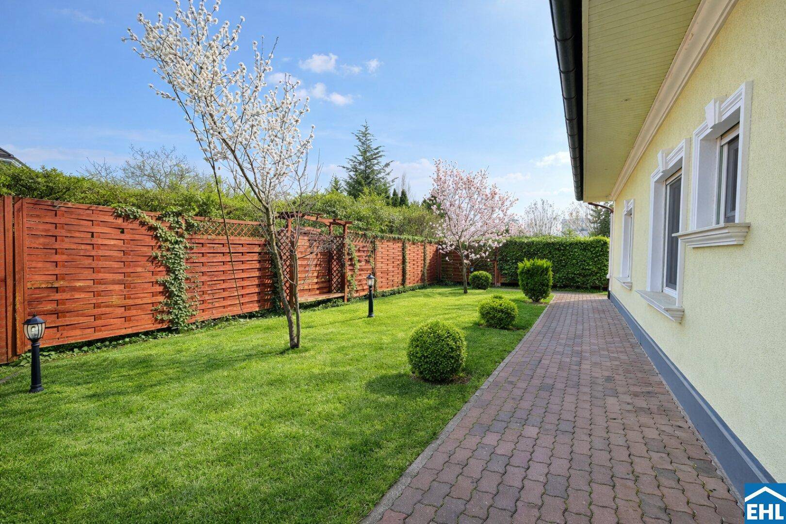 Gartenweg entlang des Hauses mit gepflegtem Rasen, blühenden Sträuchern und Holzzaun.