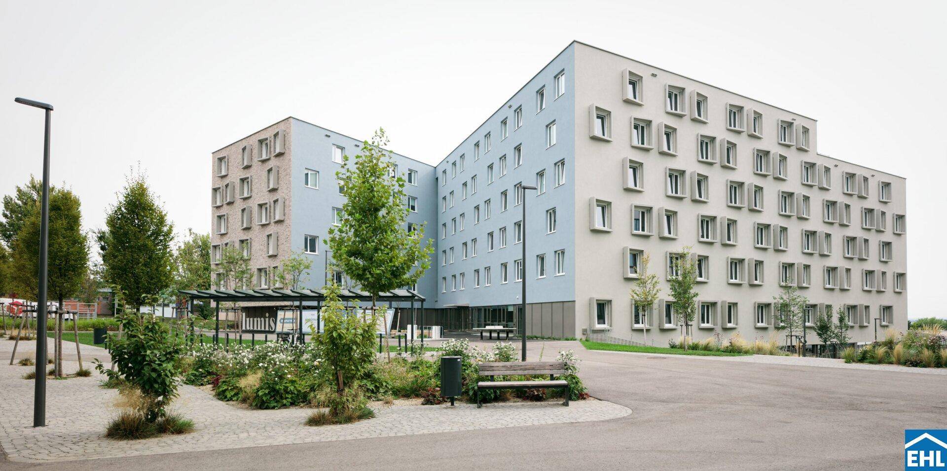 Modernes Mehrfamilienhaus mit vielen Fenstern, gepflegter Außenanlage und Sitzgelegenheiten im Grünen.
